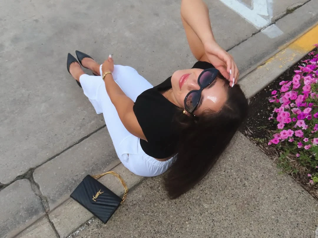 Samantha in sunglasses and a black top sitting on a curb with a designer bag
